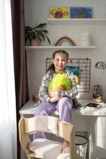 A cheerful girl sits in a sunlit room, surrounded by plush toys and colorful decor. The cozy space