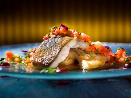 A beautifully plated gourmet hake fillet is garnished with vibrant diced tomatoes, fresh herbs, and