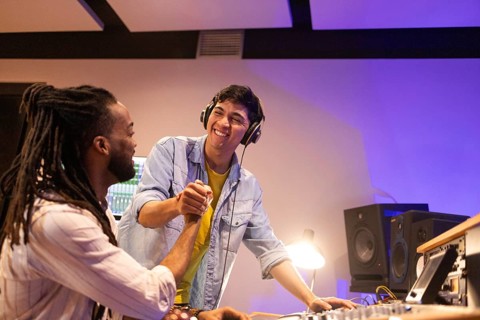 Two musicians in a recording studio share a triumphant handshake. One is seated with a sound mixer,
