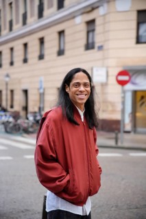 A man stands confidently on a city street, wearing a red jacket and glasses. The urban environment