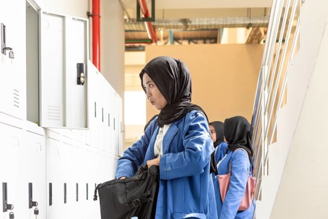 Asian Muslim student in a school hallway, wearing a blue uniform, organizing her belongings in a