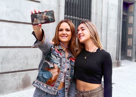 Two young women smile as they capture a selfie with a smartphone. They stand close to a building,