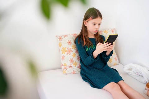 A young girl in a teal dress sits on a white bed, deeply focused on her handheld gaming console.
