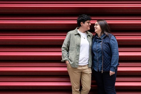 A joyful lesbian couple shares a loving moment in front of a red urban backdrop. The casual attire