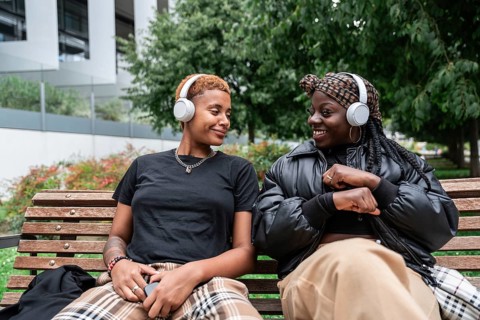 A joyful LGBT multiethnic couple listens to music with headphones, sitting on a park bench. They