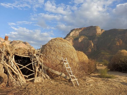 A rural village with piles of straw and mountains under a blue sky below the rock church of Abuna