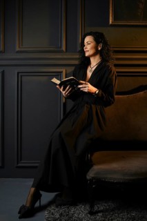A woman sits on a chair in a dark room and reads a book. The walls are dark with paneling, and