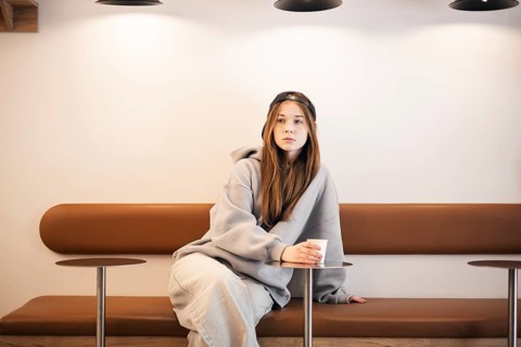 Teenage girl in casual attire with a cap, sitting thoughtfully on a bench. She holds a paper cup,