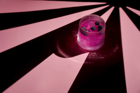 A vibrant pink drink garnished with blueberries sits in a ribbed glass, casting dramatic shadows on