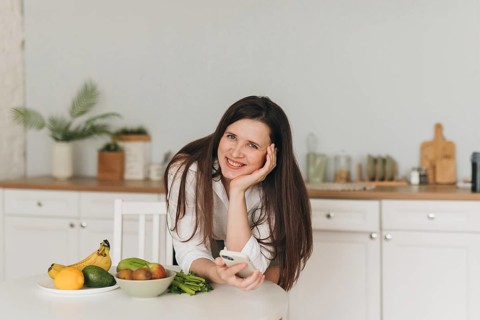A nutritionist smiles in a modern kitchen, surrounded by fresh fruits like bananas, avocados, and