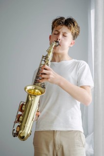 A young boy in a white shirt skillfully plays a saxophone, eyes closed in concentration. The soft