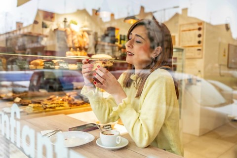 Woman savoring a flaky pastry and steaming coffee at a sunlit cafe window, enjoying a relaxed