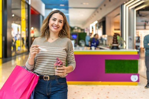 Happy young woman smiling at camera while holding shopping bags and eating ice cream in a bright