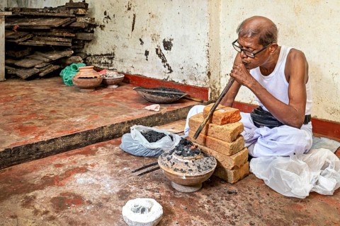 Man blows through a pipe into a coal-fired vessel in a workshop, a man burns sapphires in a small