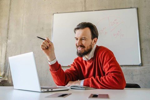 A man in a red sweater smiles while holding a pen and engaging with his laptop in a casual office