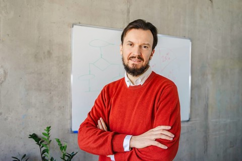 A man in a red sweater stands with arms crossed in front of a whiteboard in a modern office