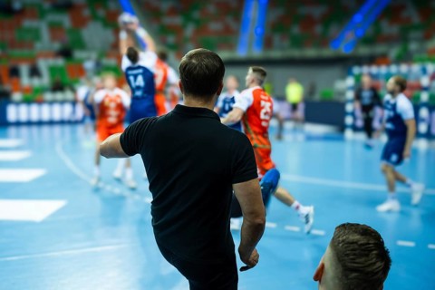 View form the behind on head coach and substitutions bench with handball match in background in