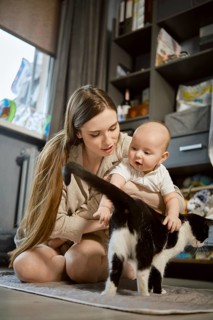 A mother kneels on the floor in a living space while her baby sits next to her. The baby looks at a