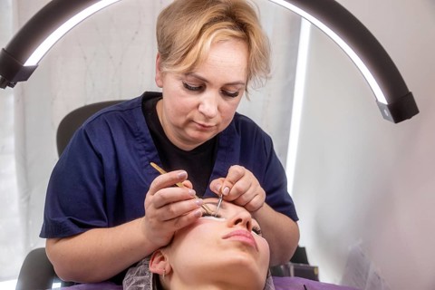 A salon technician carefully applies eyelash extensions under an LED lamp. The setting highlights
