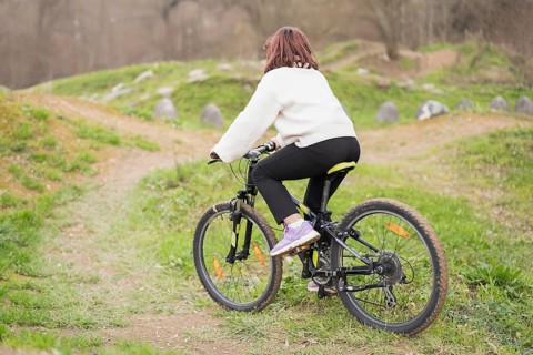 A girl wearing a white sweater and black pants rides a bicycle on a grassy, uneven trail.