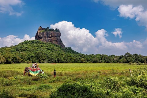 An impressive rock rises above a green plain as travelers travel on an elephant, The Lion Rock near
