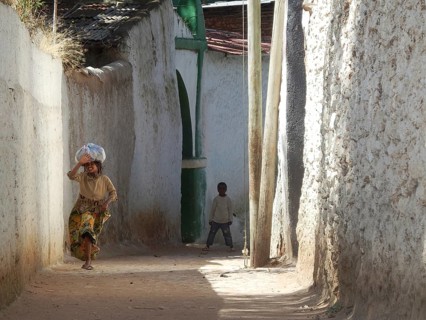 A woman carries a package on her head in a narrow alley and waves while a child watches, Harar,