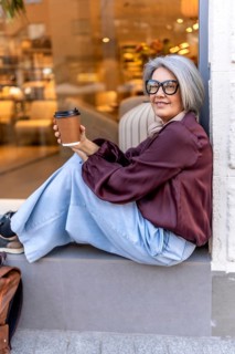 Fashionable senior woman with gray hair and eyeglasses smiling on a city curb, holding a takeaway