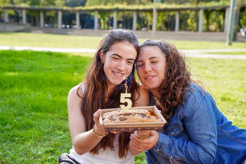 An affectionate young women couple in a park, smiling as they hold an anniversary cake with a 5