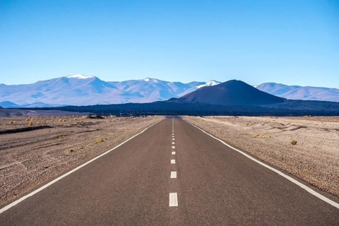 A straight road cuts through the arid desert landscape of La Puna in Argentina, with snow-capped