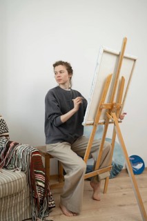A barefoot artist sitting at an easel, thoughtfully painting on a canvas. The cozy studio has a
