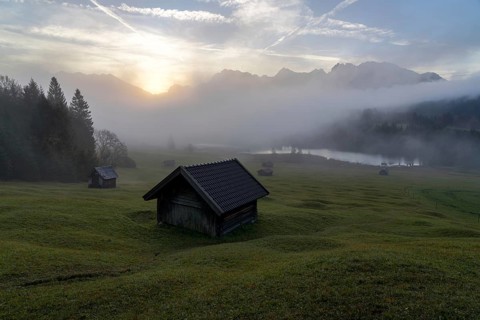 A serene view of Geroldsee in the Bavarian Alps at sunrise, featuring misty fields, rustic wooden