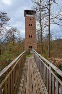 A stone tower rises majestically at the end of a wooden path surrounded by forest and dramatic sky,