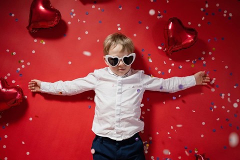 A young boy with heart-shaped sunglasses stands with outstretched arms surrounded by red