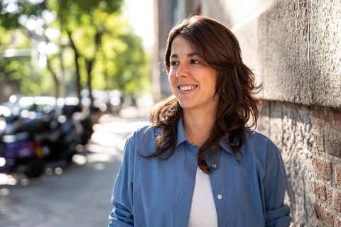 A happy lesbian woman with wavy brown hair smiles warmly while leaning against a stone wall. She is