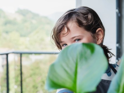 View of a young girl playfully hiding behind a large green leaf on a balcony, looking at camera,