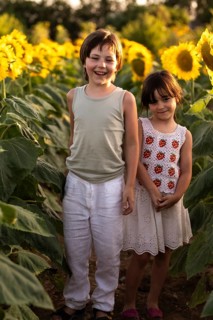 Two children stand joyfully in a sunflower field during golden hour. The warm sunlight highlights