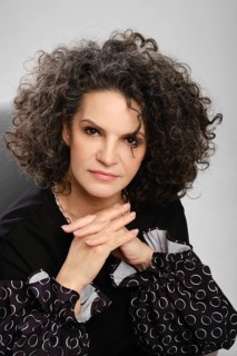 Closeup portrait of senior woman with lush curly hair and black pantsuit posing in studio over grey