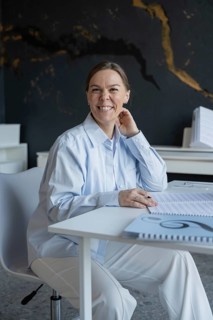 A smiling woman sits at a desk preparing for a piano lesson, surrounded by sheet music and notes,