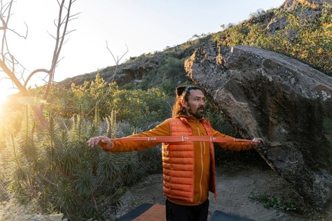 A climber stands in front of a large boulder in a natural setting, stretching with a resistance