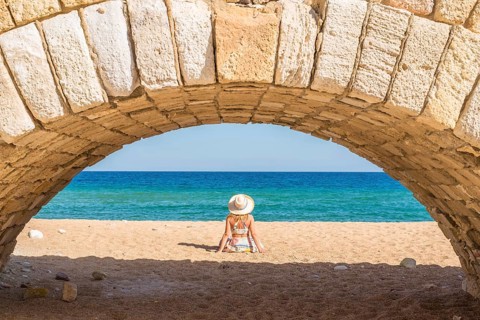 A woman enjoys a peaceful moment on a sandy beach under a stone arch, with the vibrant blue sea