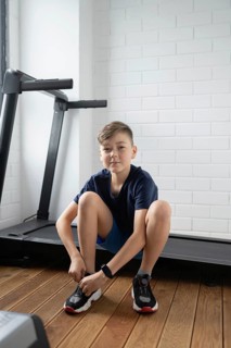 Young boy tying his shoes in a modern home gym with a treadmill in the background. He wears a smart