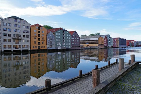 Colourful buildings are reflected in the calm waters of a river next to a wooden dock, Nidelva,