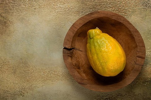 A lemon with a bright yellow hue sits in a rustic wooden bowl against a muted background,