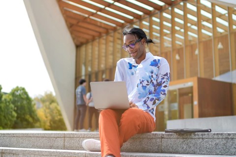 A man in a floral shirt and red pants sits outside, working on a laptop. The scene exudes