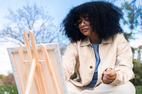A young black artist with curly hair paints on a canvas in an outdoor setting. Wearing glasses and