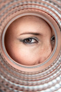 A close-up of a woman's eye peering through the circular opening of a textured pipe, capturing the