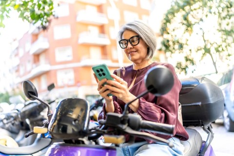 Senior gray haired woman with glasses sitting on a scooter on a sunny city street, smiling as she
