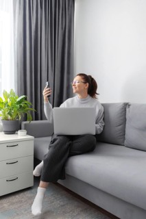 A woman sitting on a comfortable gray sofa, holding a phone and using a laptop for work in a cozy