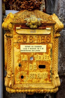 The yellow mailbox, aged and textured, stands against a rugged wall, featuring classic designs and