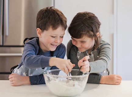 Two joyful siblings mix ingredients in a glass bowl, wearing aprons in a well lit kitchen. Their